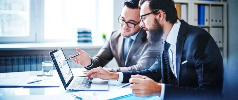 Dois homens brancos em um escritório olhando um computador e discutindo algum assunto.