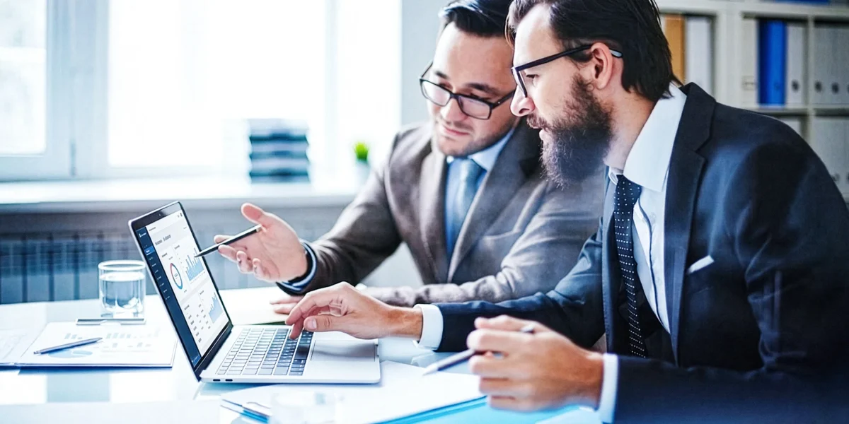 Dois homens brancos em um escritório olhando um computador e discutindo algum assunto.